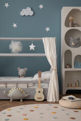 Aesthetic composition of cozy kid room interior with stylish bed, blue wall, braided pouf, round rug, books, colorful garland, gray curtain and personal accessories. Home decor. Template.