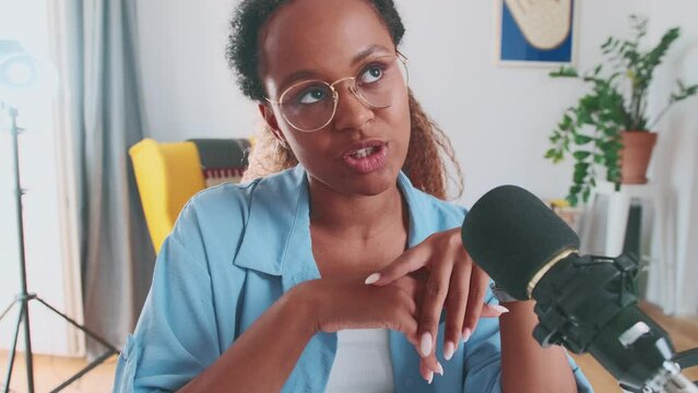 Young sensible attractive African American woman talking and looking at camera sits near professional microphone located in home studio for blogging. Blogger, influencer, journalist