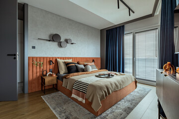 Interior design of elegant bedroom with big orange bed, beige and grey bedclothes, blue curtain, rug, modern lamp, night stand, vase with dried flowers and personal accessories. Home decor. Template.
