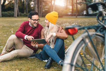 Smiling confident man and woman using digital tablet pc while sitting on grass in public park. Well dressed attractive people relaxing with tablet device in autumn.