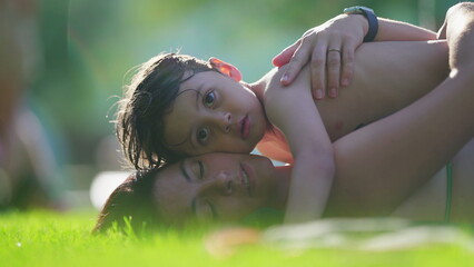 Affectionate moment of little son kissing mother in cheek while laid on grass during summer day...