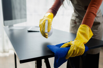 Cleaning desk surface in office with sanitizer spray, wear gloves and wipe the table with a towel, the housekeeper is cleaning the work desk for hygiene because of the Covid-19, cleaning idea. - obrazy, fototapety, plakaty