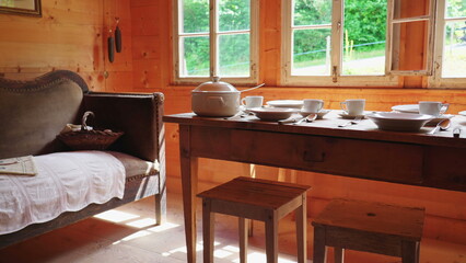 Rustic setting of wooden table and window at rural chalet, antique couch with white dinnerware. Humble ceramic plates, teacups, and utensils on aged table