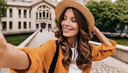 Young happy woman wearing hat winking taking selfie on summer city street. Cheerful pretty lady fashion model holding phone looking at mobile camera making portrait photo outdoor