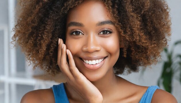 Stunning happy African American black young woman with curly afro hair and perfect smile looking at camera touching face with hand. Salon skincare, cosmetology and spa concept. Close up. Copy space.