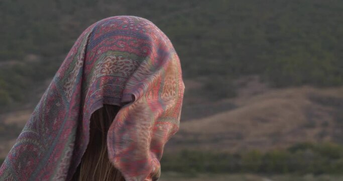 A lonely woman with a scarf on her face against the backdrop of a mountain landscape at sunset. The wind tries to blow away the scarf and free the personality. Visual display of psychological state