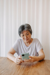 Happy asian old elder woman smiling and looking at mobile, holding and using phone, chatting or reading news, sitting on chair alone over white curtain at cafe with coffee and croissant on wood table.