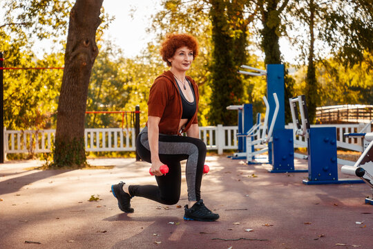 Adult Caucasian pretty woman is doing sports in park on street. Athlete does squats with dumbbells in her hands. Healthy lifestyle