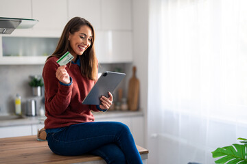 Happy young businesswoman holding credit card and looking at digital tablet pc while sitting on kitchen counter at home.