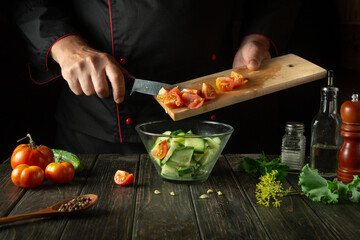 The cook is preparing a vegetable salad in the kitchen. The concept of cooking vitamin salad with tomatoes and cucumbers. Space for a recipe on a dark background