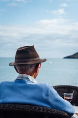 Fototapeta premium Homme avec un verre regardant la mer. Homme assis en terrasse sur la plage. Se relaxer à la mer. Prendre des vacances. Retraité dans le Sud. Sénior en vacances. Apéritif à la mer. Regarder l'horizon
