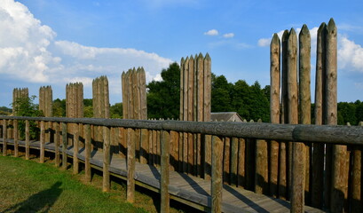 A close up on an old borough or village surrounded by a wooden palisade made out of logs, planks, and boards and a small moat below the hill seen on a sunny summer day next to some forests and moors