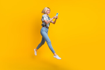 Fototapeta na wymiar Full length photo of optimistic clever girl dressed print shirt jumping read email on smartphone isolated on yellow color background