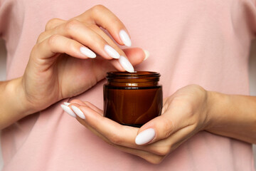 A young woman holds a brown glass jar with moisturizing cream in her beautiful well-groomed hands. Hand and body skin care concept. A massage candle against a dusty rose T-shirt background. Close-up.