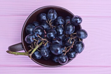fresh blue ripe grapes on a branch in a brown ceramic cup stands on a pink wooden table