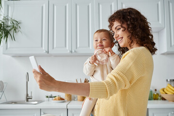 excited mother taking selfie on smartphone with toddler child holding baby bottle, candid moment