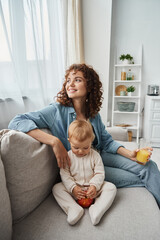 delighted woman with orange juice looking away on couch near adorable child with apple, happiness