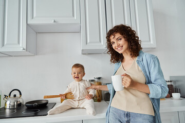 woman with coffee cup smiling at camera near baby girl sitting on kitchen counter with wooden fork