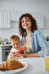 joyful mother looking at camera during breakfast with baby girl near ripe apple and tasty croissant