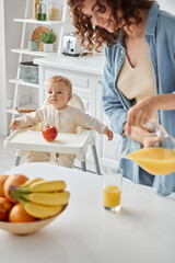 Obraz na płótnie Canvas cute child sitting in baby chair near apple while happy mom pouring fresh orange juice for breakfast