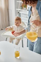 woman with jug of orange juice pointing at ripe apple near little daughter sitting in baby chair