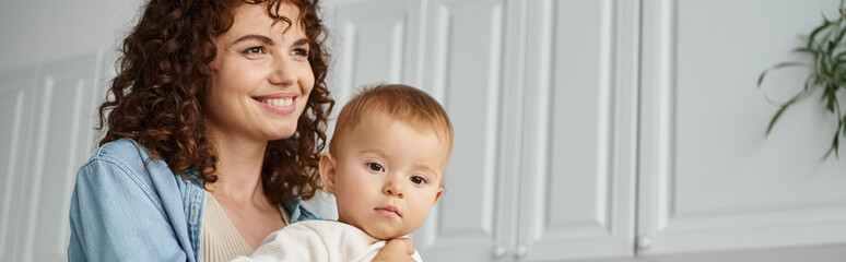 happy woman with wavy hair smiling and looking away near adorable baby girl in kitchen, banner