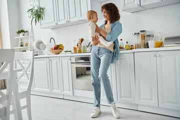 full length of happy woman holding little daughter in spacious modern kitchen with white furniture