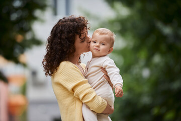 Fototapeta premium woman with wavy hair embracing and kissing cute little daughter outdoors, blissful motherhood