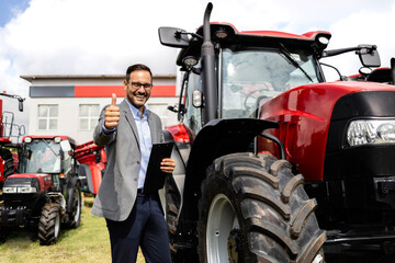 Portrait of successful tractor dealer standing by farming equipment and holding thumbs up. - obrazy, fototapety, plakaty