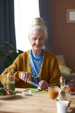 Vertical image of senior woman cutting vegetables for salad while sitting at table in the room