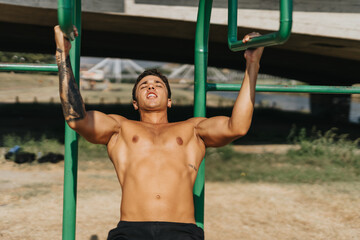 Fit male doing pull ups on metal bars in a sunny park. His dedication and persistence towards a healthy lifestyle is evident as he engage in outdoor exercise and training.