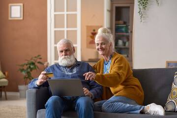 Senior couple sitting on sofa with laptop in the living room and using credit card to pay for...
