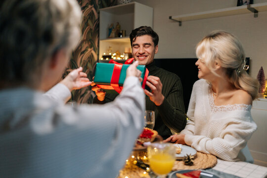Back view of unrecognizable mother-in-law giving festive gift box with Christmas present to happy man sitting with family at dinner feast table at home on xmas eve. Concept of home festive atmosphere.