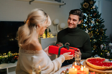 Happy surprised young woman receiving gift box from loving man sitting at dinner table at home on Christmas Eve near decorated xmas tree. Caucasian male giving present to beloved female on holidays.