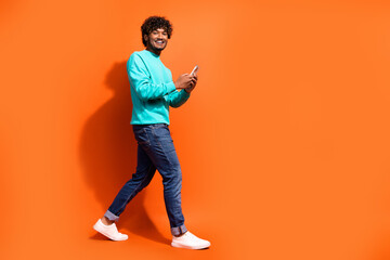 Fototapeta na wymiar Side profile shot of young arab guy going to a date with smartphone wearing stylish casual clothing isolated on orange color background