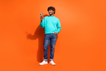 Full length photo of optimistic indian guy browsing social media smartphone while waiting his coffee isolated on orange color background