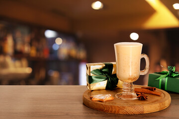 Glass of delicious eggnog, spices, cookie and gift boxes on wooden table in bar, space for text