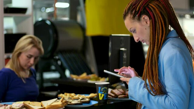 a woman with red dreadlocks works on the phone during a corporate party, food is on the table in the office, two young girls are finishing work in a blue sweater. a blonde is typing on computer