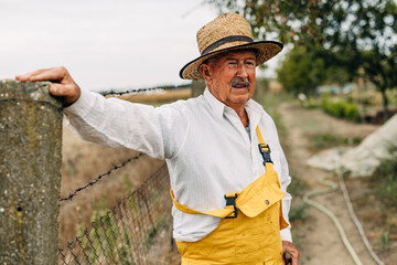 Old man leaning on a fence of his garden and pensive looking away.