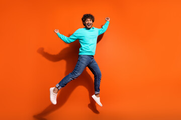 Full length photo of sportive indian guy curls hair wear denim jeans with cyan pullover win lottery isolated on orange color background