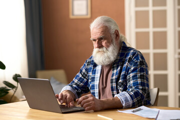 Serious senior man typing on laptop while sitting at table in the room
