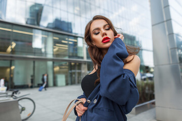 Stylish beautiful elegant fashionable woman with red lips with a hairstyle in a fashion blue suit walks on the street near the office building