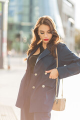 Obraz premium Beautiful fashion urban female portrait of a pretty stylish elegant business girl in a fashionable blue suit with a bag walks in the city on a sunny day. Trendy lady