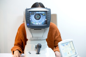 Woman looking through auto refractor machine during an eye examination in a clinic, displaying eye...