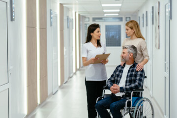 Friendly female doctor with clipboard talking to a female, her husband patient sitting on a wheelchair and her partner all looking happy.