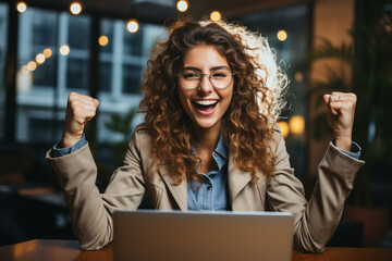 Joyful woman with curly hair celebrating success at a laptop in a cafe