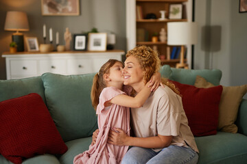 Cute little girl kissing her mother on sofa in living room