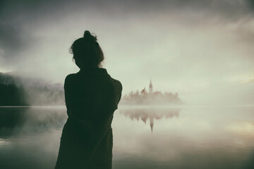 Woman in gray coat watching a sunrise among the fog on the shores of Lake Bled, Slovenia III