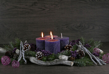 Adventsdekoration: Adventskranz mit drei lila brennenden Kerzen für den dritten Advent mit  Tannenzweigen und Weihnachtsdeko vor einer Holzwand mit Platz für Text.