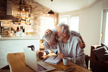 Happy senior couple writing on paper with laptop at home - obrazy, fototapety, plakaty